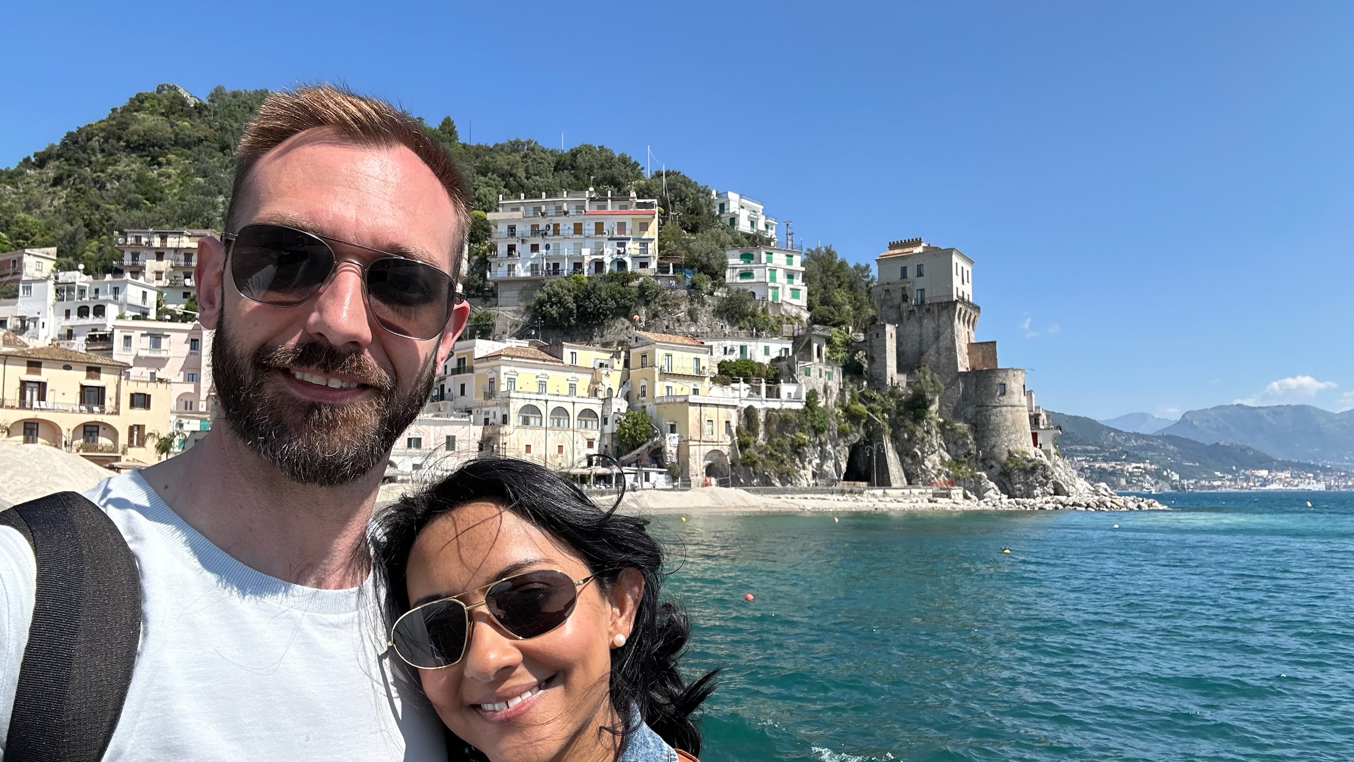 Kari and Anton selfie with a hillside coastal town and turquoise sea behind them